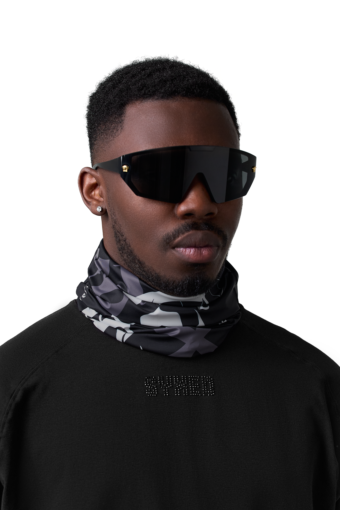 A man wearing oversized black shield-style sunglasses and a gray, black, and white camouflage neck gaiter, set against a plain white background.