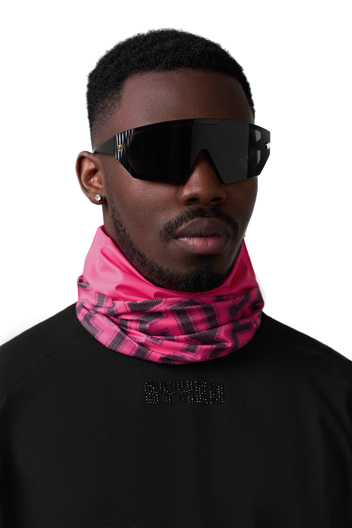 A man wearing a bright pink and black plaid neck gaiter, black shield-style sunglasses, and a black sweatshirt against a plain white background.