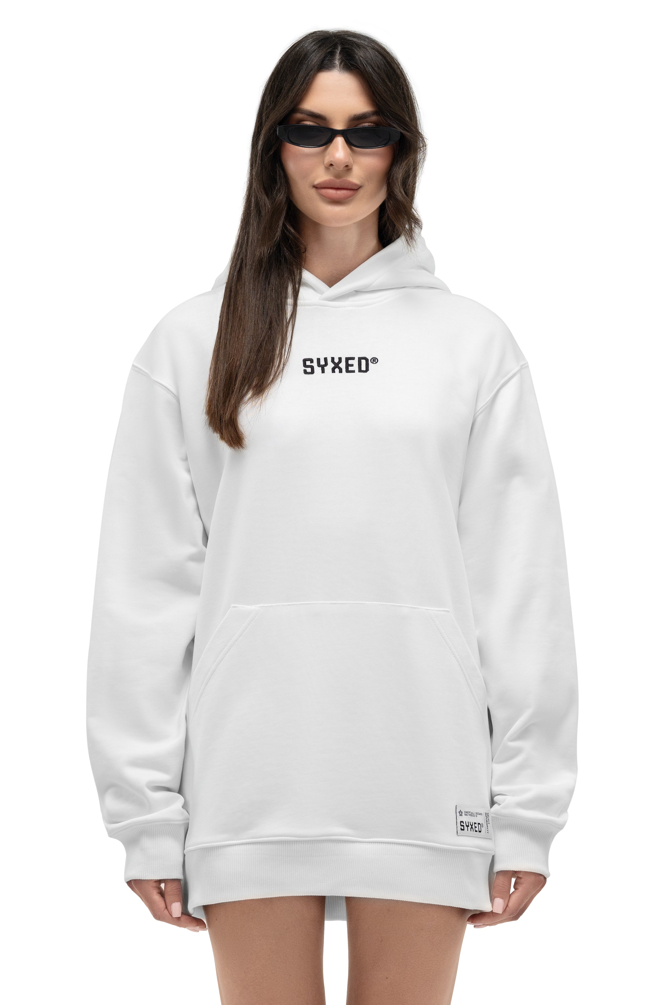 A model wearing an oversized white hoodie with 'SYXED' printed in black across the chest, styled with black sunglasses against a plain white background.