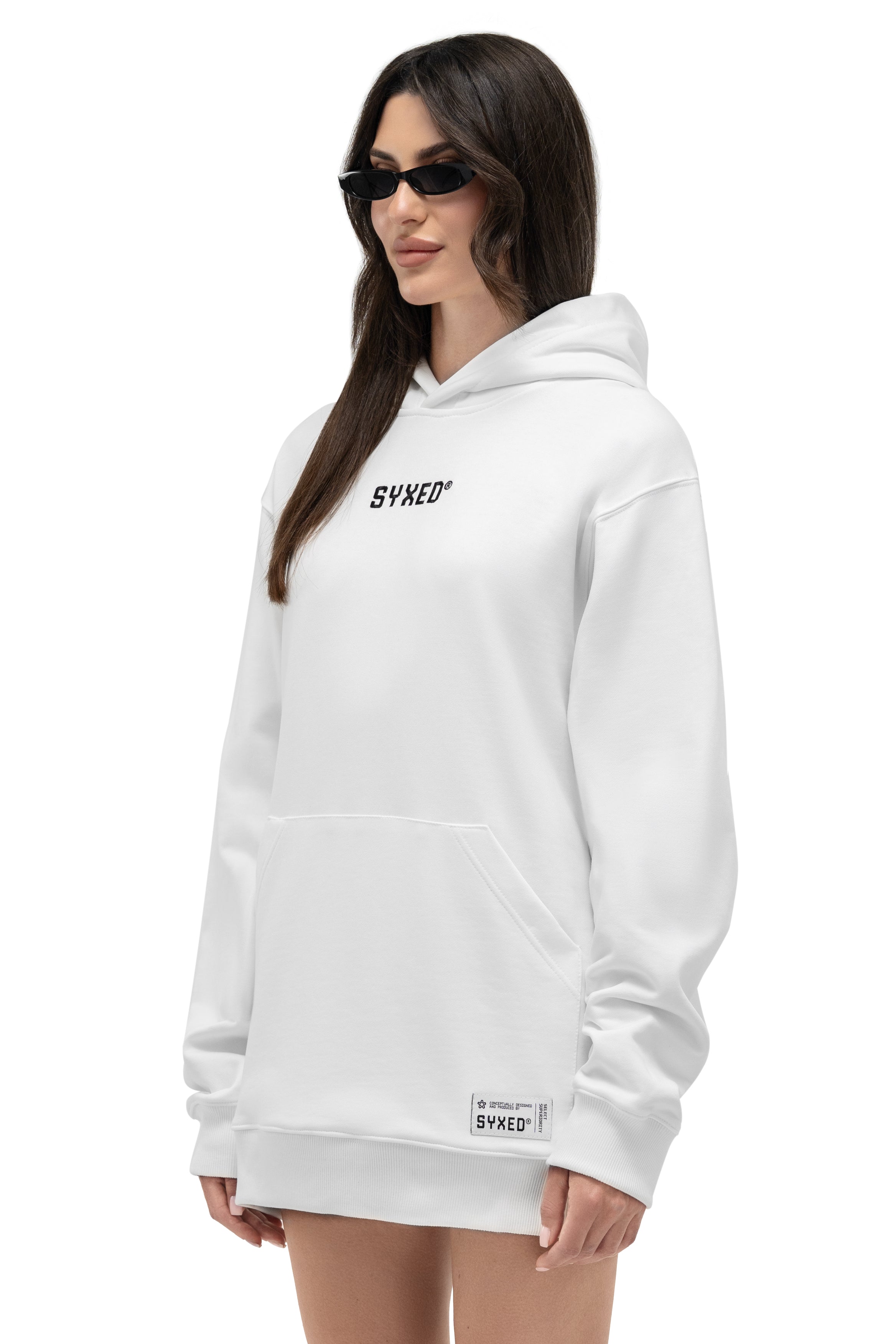 A woman modeling a white pullover hoodie with a black 'SYXED' logo on the chest and a small brand patch near the hem, photographed against a plain white background.