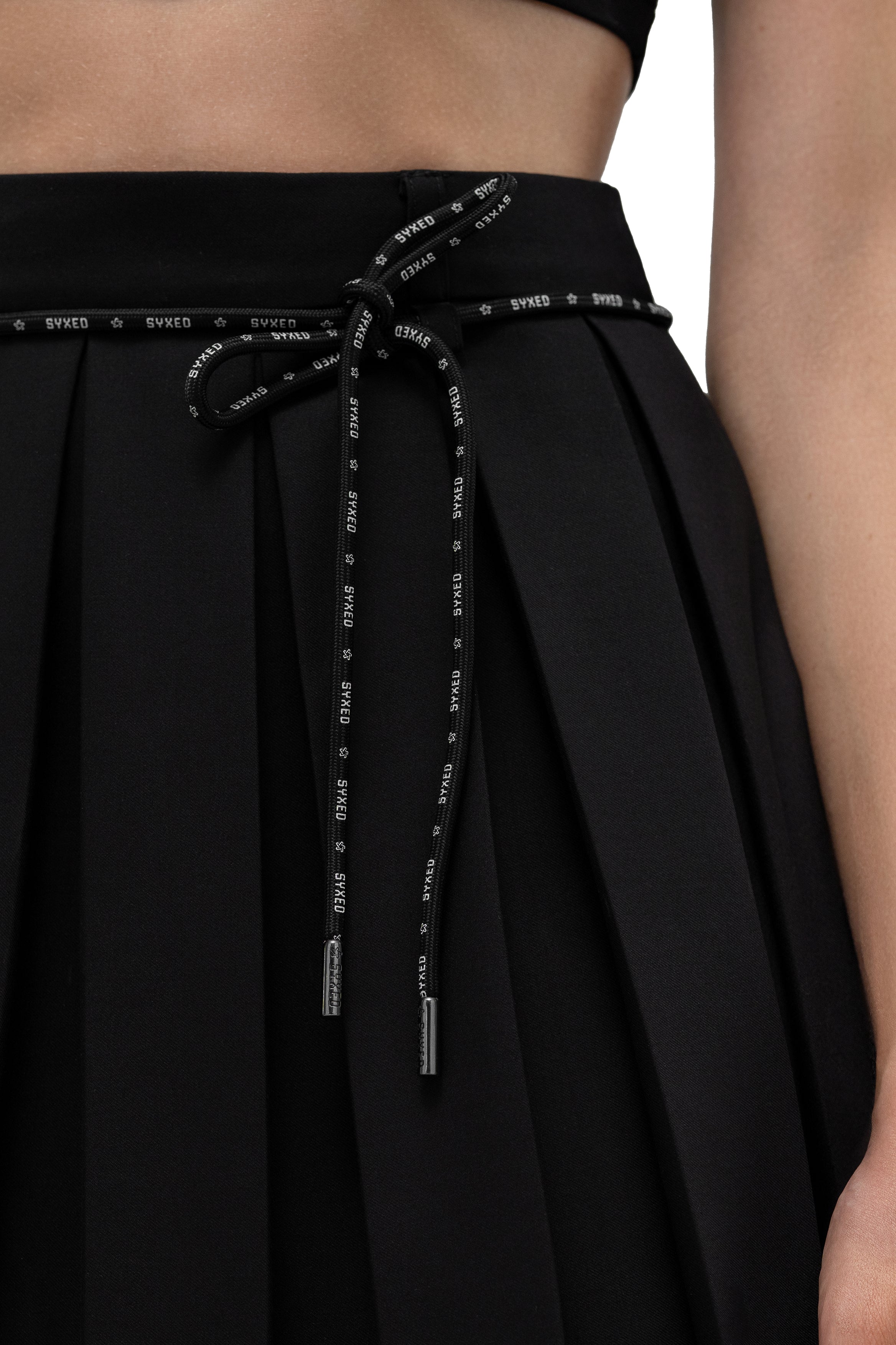 Close-up of a black pleated skirt with a matching waistband and drawstring tie. The drawstring is black with white 'SWEET' text repeated along it, and the waistband also has small white text details.