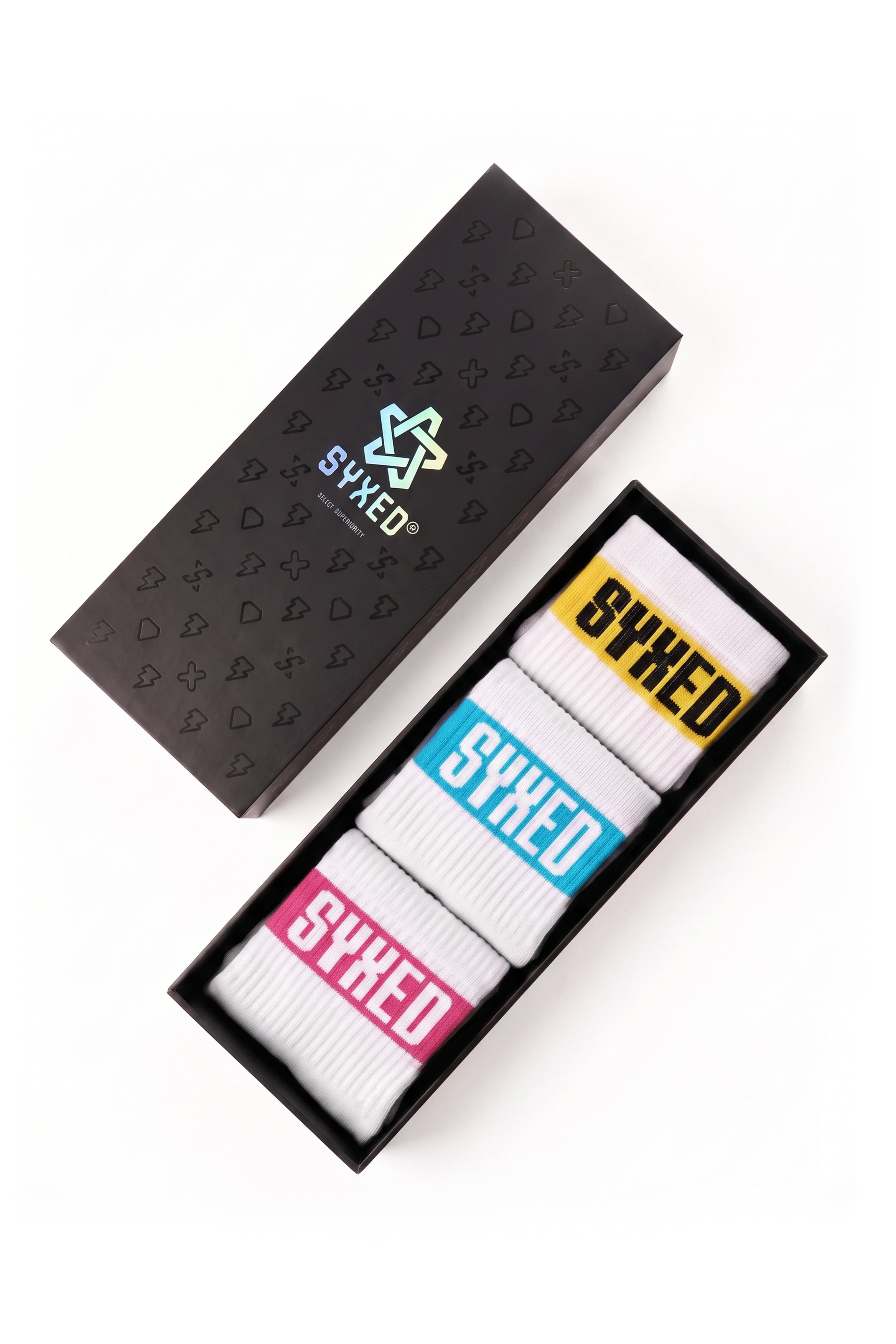 Three pairs of white crew socks in a black presentation box, each with a different colored 'SYXED' label in yellow, blue, and pink, next to the black branded outer box on a white background.