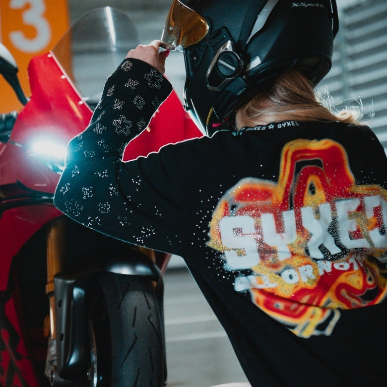Person wearing a black jacket with colorful graphics and text, standing next to a red motorcycle.