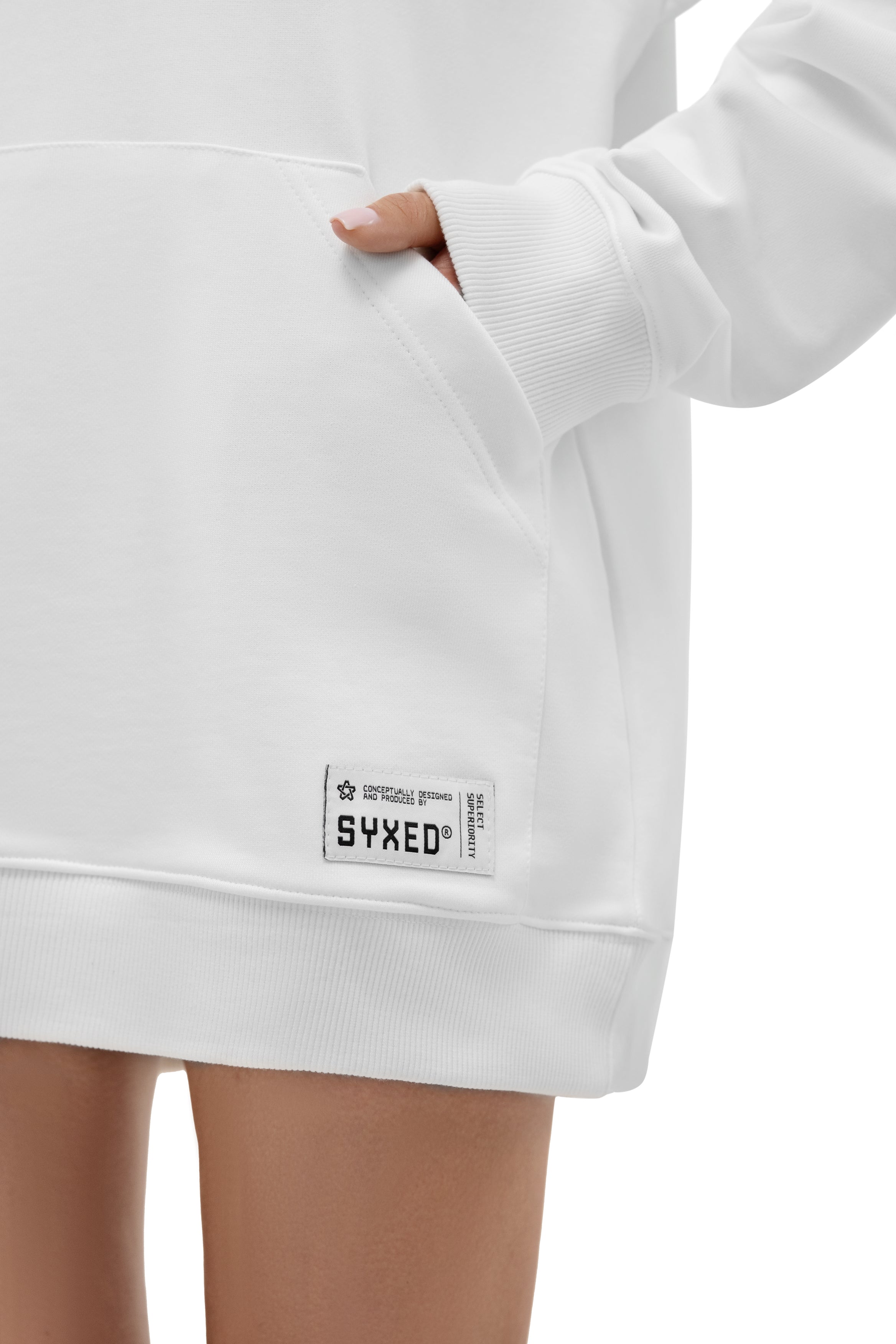 Close-up of a white pullover hoodie worn by a model, showing the front pocket with a hand inside, ribbed cuffs and hem, and a small white brand patch reading SYXED near the lower left front.