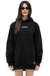 A woman modeling an oversized black hoodie with 'SYXED' printed in white across the chest and a small matching logo patch near the lower hem, shot against a plain white background.